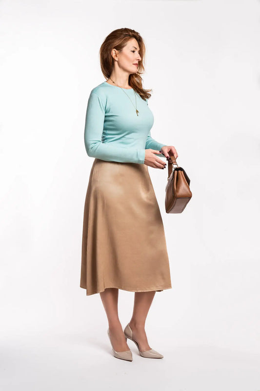 Woman wearing a light blue merino blouse and beige skirt holding a brown handbag on a white background