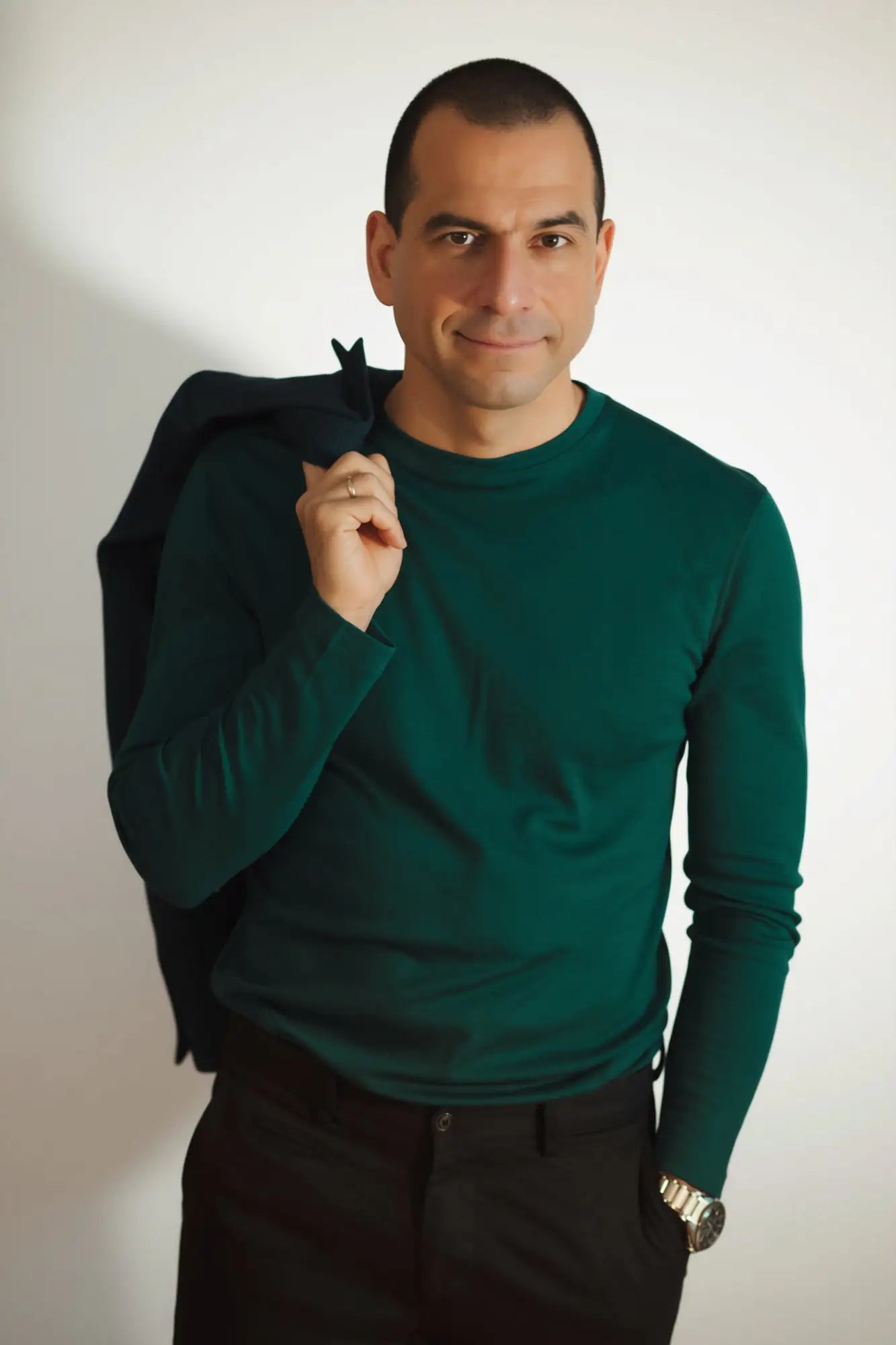 Man wearing a green merino top holding a black jacket against a white background
