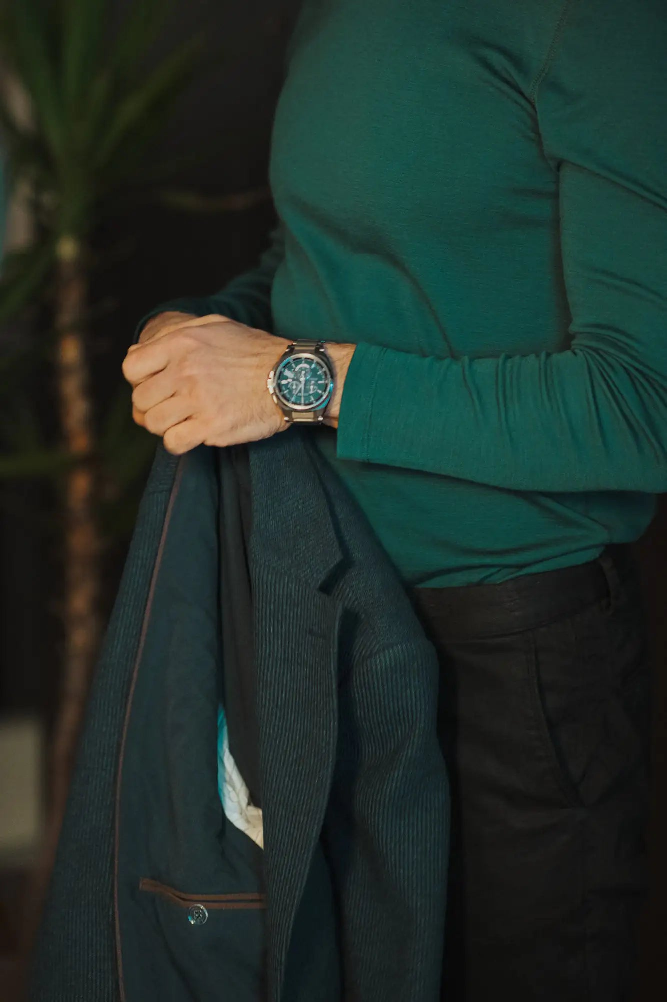 Person wearing a green merino top and dark pants with a watch, holding a dark jacket.