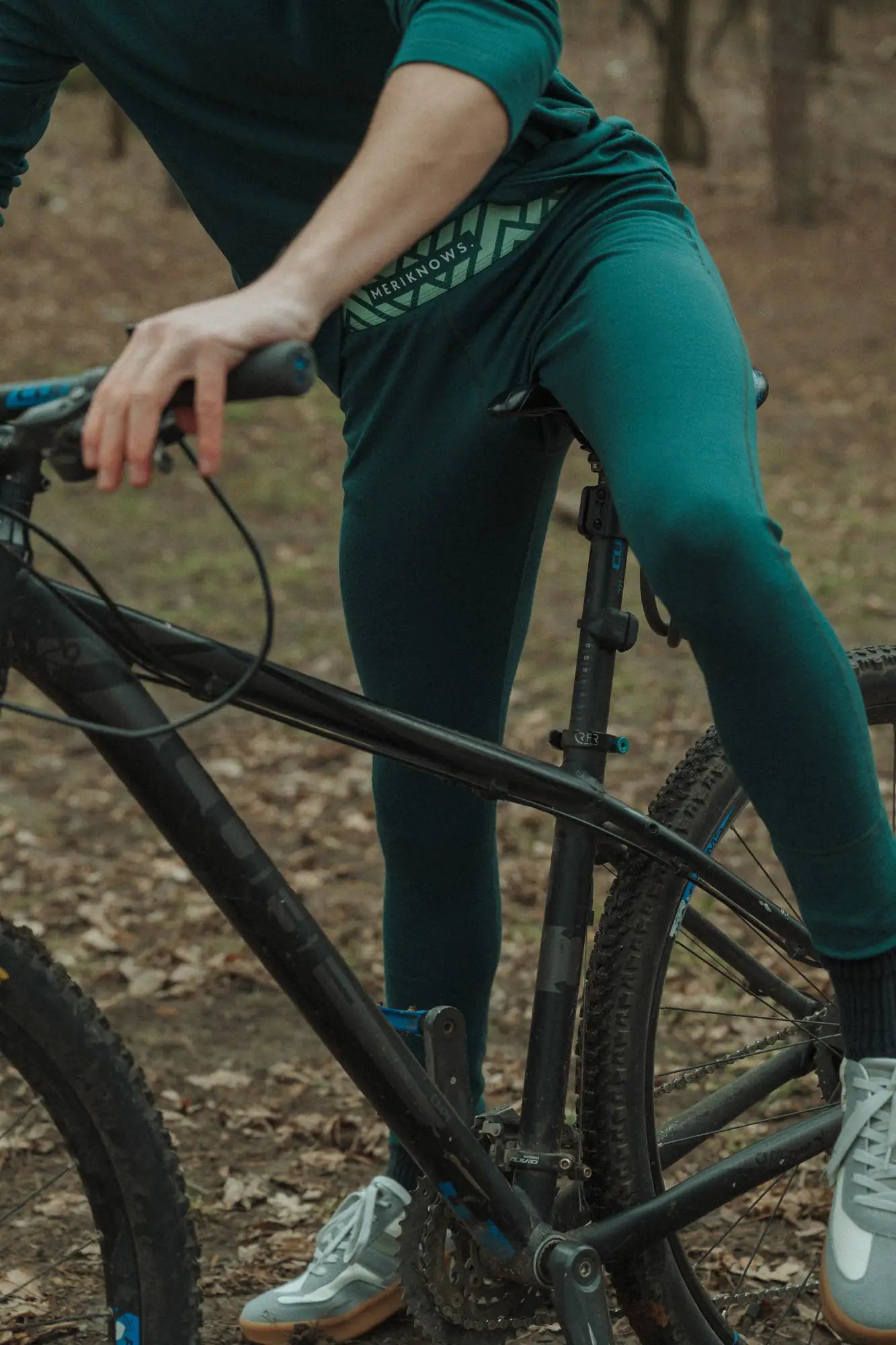 Person wearing green merino wool pants with a brand logo, standing next to a bicycle in a forest setting.