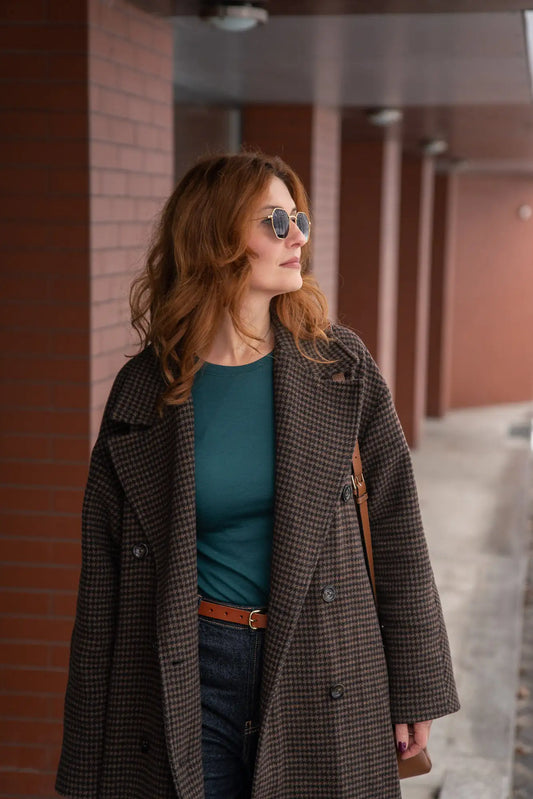 Woman wearing a fine merino long sleeve blouse, checkered coat and sunglasses, standing against a brick wall.