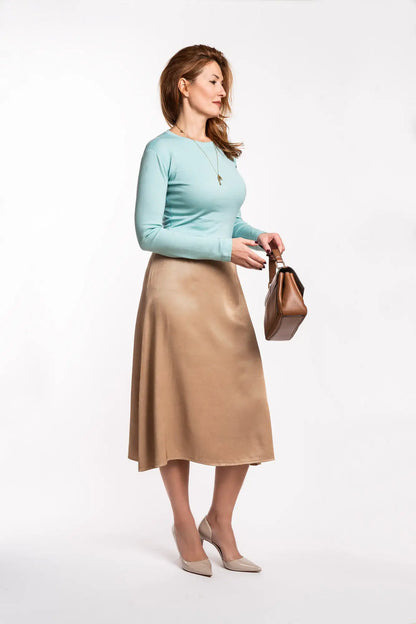 Woman wearing a light blue merino blouse and beige skirt holding a brown handbag on a white background